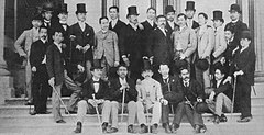 Photo of a large group of men on steps. Some are seated, and others are standing; several are wearing top hats.