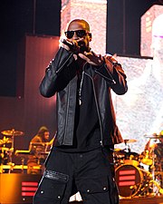 A black man wearing sunglasses and a dark outfit performs with a microphone