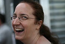 Photograph of Jeni Tennison. She is looking back over her shoulder toward the camera laughing. She is a white female with long dark hair, tied back, and is wearing glasses.