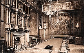 An elaborately-furnished room with a large fireplace, tapestry on the wall, and table and chairs in the centre