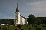 Bild einer weißen Holzkirche mit dunklem Dach. Im Vordergrund befindet sich ein Friedhof