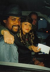 A man and a woman smile to the camera while seated in a car. They both wear black hats and the car's driver can be seen smiling in the background too.