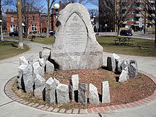 circular monument in a park, and made of multiple grey stones. The large central stone contains a bilingual inscription in memory of women killed by men's violence. Many much smaller irregularly shaped stone shafts are carved with women's names