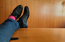 Gambe che indossano dei pantaloni e scarpe scuri, ma calzini di colore diverso: uno nero, uno fucsia. Le gambe sono appoggiate sul bordo di un tavolo di un ufficio.