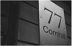 Sur un mur apparaît une plaque qui porte « 77 Cornhill ».