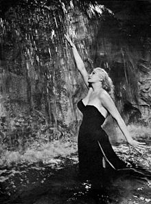 Une jeune femme en robe de soirée dans une fontaine tend un bras dans l'eau qui coule et sourit - c'est Anita Ekberg