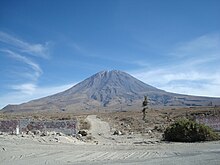 Picture of small brown mountain, taken from ground level, with a dirt road leading up to it.