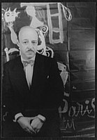 Portrait of writer Charles R. Jackson by photographer Carl Van Vechten. Jackson is facing the camera. He is partly bald and has a pencil moustache. He is wearing a dark suit with a white shirt, a white handkerchief, and a polka-dot bow-tie. In the background is a dark brick wall with white art glyphs.
