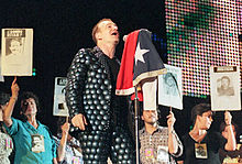 A light-skinned man with brown hair singing into a microphone on a stand, which has a flag draped over it. His shirt and trousers are both grey and feature a design of many overlapping circles. He faces to the right. A line of women stand behind him, each one holding up a sign that says "Donde Estan" or "Justcia". Every sign has an image of a different person below the text.