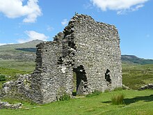 The west tower of the castle, looking north. The tower is craggy and ruined, and a mountain is visible in the distance.