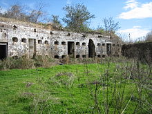 Mur en brique grise avec plusieurs portes et fenêtres. L'endroit semble abandonné et est recouvert de mauvaises herbes.