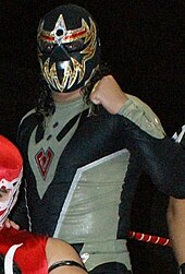 A masked professional wrestler in the ring, wearing a black mask and black bodysuit, both with gold markings