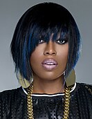 Missy Elliott posing on a grey background wearing a black shirt and gold jewelry.