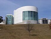 A large white and cement building sits on top of a large hill. A large round part of the building jets out from the rest and contains many windows.