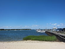 view from sandy shoreline out into the water with moored sailboat in the distance and the wharf on the right