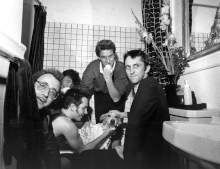 Five males in a bathroom surrounding a small dog that has shampoo spread on its fur.