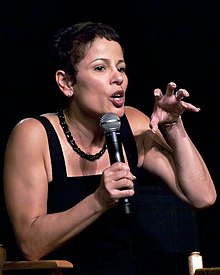 A woman with short brown hair and a black top is talking into a microphone while gesturing with her left hand.