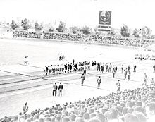 Photo en noir et blanc d'un avant-match de football, prise depuis les tribunes, avec le terrain au second plan