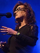 A dark-haired woman wearing a dark blouse and glasses sings into a microphone.