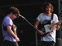 Two guitarists are performing a song live on a stage, in front of a mic mounted on a microphone stand.