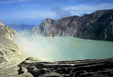 A photograph depicting a blue sky with white clouds at the top, a light grey lake in the middle, and dark grey rocks surrounding the lake.