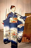 A young woman stood up in a dark blue long-sleeved kimono with a white designs across the lap and the middle of the sleeves. The sash she wears is orange with a thin green belt in the centre