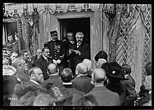 Photographie noir et blanc d'un homme moustachu lisant un discours à l'entrée d'une maison, devant un public d'hommes et de femmes.