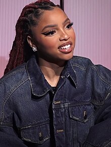 A woman with long, thick, dark hair behind her wears a blue denim jacket and smiles.