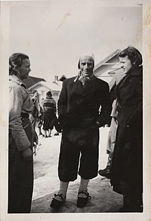 Black and white image of Per Samuelshaug at approximately 33 years old. He is stood in the snow with two women. He is wearing a black coat and black trousers with a white hat and white socks