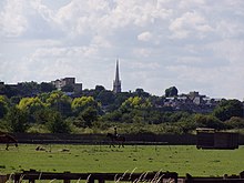 A wooded urban landscape with a church tower as one of the buildings emerging above the trees. The tower is surmounted by a spire that rises well above all other buildings on the hillside.