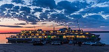 Photographie d'un bateau touristique à quai au couchant sur le Ienisseï. Les lumières du bateau sont allumées, mais le soleil n'est pas encore couché.