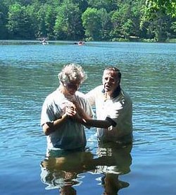 A baptism in a river. Two men are standing hip-deep in blue water with trees in the background.
