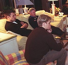 Bill Clinton, Ben Affleck, and Matt Damon sit on two sofas while looking towards a television screen