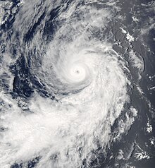 A photograph of a hurricane over the Eastern Pacific Ocean. It has a well-defined eye embedded by a round area of thick clouds, though most of the hurricane's cloud cover is located in a long, wide band that curves around the right and bottom flanks.