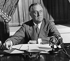 Photograph portrait of Franklin D. Roosevelt sitting at a desk with sheets of paper and a microphone in front of him. He wears a collared shirt, a necktie, and a suit coat.