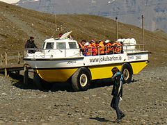 "Véhicule jaune et blanc a l’arrêt au sec devant un ponton, avec une vingtaine de personnes à bord portant des gilets de sécurité, dans un paysage minéral"