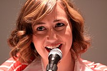 In a gray background, a young woman with a microphone in front of her smiles greatly.