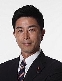 Japanese man, clean shaven, smiling slightly with with closed lips, has coiffed possibly gelled back black hair, pictured framed as shoulders and head, wearing a suit with white shirt and black coat, tie is navy blue with a pink or salmon somewhat shiny thick diagonal stripe design. Wearing a round lapel pin on his left lapel with unclear detail as well as what looks to probably be a lavalier microphone