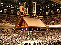 Inside of the arena during a tournament, the place is crowded. The Japanese flag is seen on top of the Shinto roof and portraits of previous winners are hang