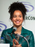 A mid shot of a young woman with tied up curly brown hair, wearing a green jacket. She is talking into a microphone at a press event and smiling at the audience.