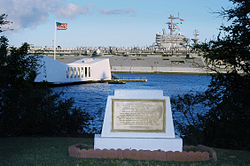 A golden plaque on stone on the shore of Ford Island, with a white memorial bridge floating over the USS Arizona