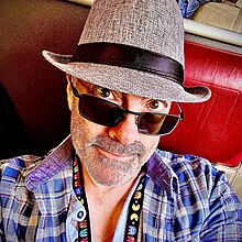 The musician Tim Wright also knows as CoLD SToRAGE sitting in a Swiss train wearing a trilby hat and looking up into the camera over the brim of his sunglasses, wearing a check-shirt and sat on the train's bright red seat. Photo was taken of himseld using his iPhone 14.