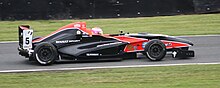 Photographie d'une monoplace de Formule Renault noire et rouge vue de profil droit, sur une piste, avec une pilote au casque rose.
