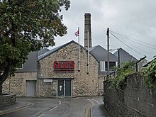The front of an old stone building with GILKES in large red letters and the royal coat of arms, flying the union flag from a flagpole. The entrance door is modern. An old stone industrial chimney and a series of modern roofs are seen beyond.