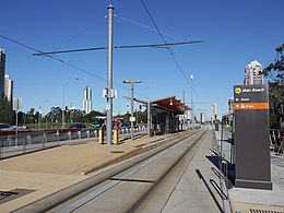 Island platform with a canopy in the centre. A shallow ramp leads up to the platform. At the top of the ramp an overhead wiring support mast is flanked by ticket validation machines on either side. A sign at the entrance to the stop directs passenger to buses or the beach.