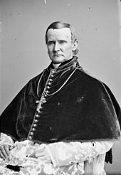 A seated man wearing liturgical vestments and pectoral cross faces forward.