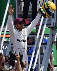 Photo of Lewis Hamilton wearing a red cap and silver overalls holding his arms aloft with a yellow helmet in his left hand