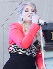 A blonde woman wearing a hot pink cardigan over a leopard print top and a black skirt singing behind a microphone stand on stage