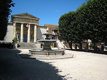 Photographie en couleurs d'une place dont une fontaine occupe le centre ; bâtiment à colonnes et fronton triangulaire en arrière-plan.