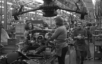 Black and white photo of 2 women at an assembly machine. A migrant worker walks past them, wearing a VW branded apron